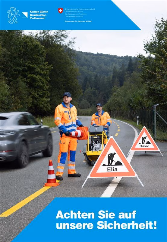 Zwei Strassenarbeiter in orangefarbener Arbeitskleidung sichern eine Fahrbahn mit Verkehrsleitkegeln und dreieckigen Warnschildern, die mit den Namen «Elia» und «David» beschriftet sind. Ein graues Auto fährt vorbei. Im Hintergrund Bäume und ein bewaldeter Hang. Der Text im Vordergrund lautet: «Achten Sie auf unsere Sicherheit!».
